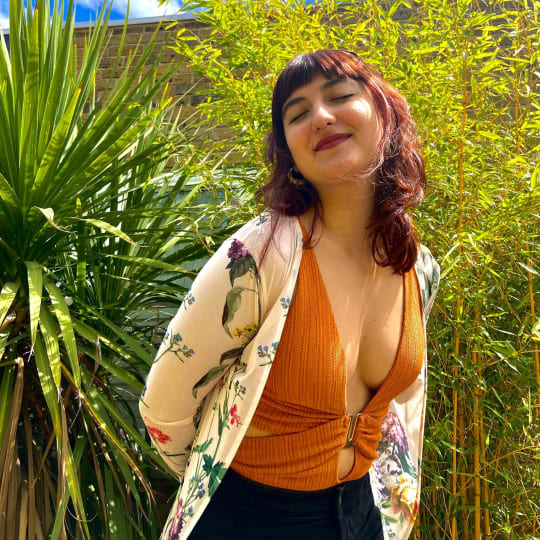 Girl with red hair smiling in an orange swimsuit and floral cardigan, in front of some green plants