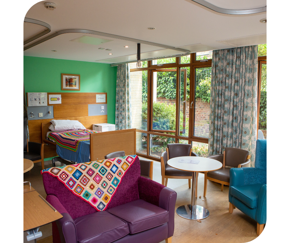 A room with a hospital bed, a purple sofa, tables and chairs with handmade crotchet blankets with a garden outside