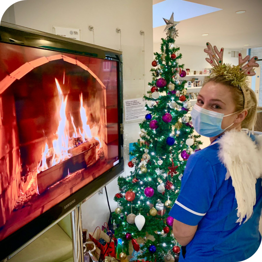 Female nurse standing in front of a Christmas tree wearing a mask, reindeer antlers, fairy wings, next to a fire on a tv screen
